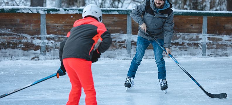Zwei Personen spielen Eishockey auf einem gefrorenen Teich. Einer trägt eine rote Hose und der andere eine Mütze. Bäume sind im Hintergrund zu sehen.