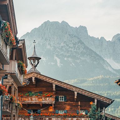 Traditionelles Tiroler Holzhaus mit blumengeschmücktem Balkon vor imposantem Bergmassiv, an einem klaren Tag, im Hintergrund wolkenverhangene Gipfel.