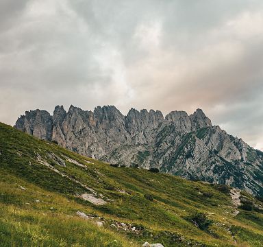 Imposantes Bergmassiv mit steilen, gezackten Gipfeln unter teils bedecktem Himmel. Im Vordergrund grasbewachsene Hügel, die sanft ansteigen.