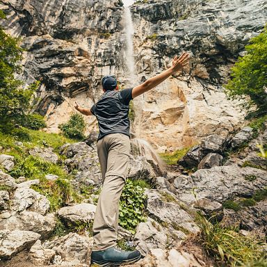 Ein Mensch steht mit ausgestreckten Armen vor einem Wasserfall, umgeben von Felsen und dichter Vegetation, und blickt nach oben zur Quelle.