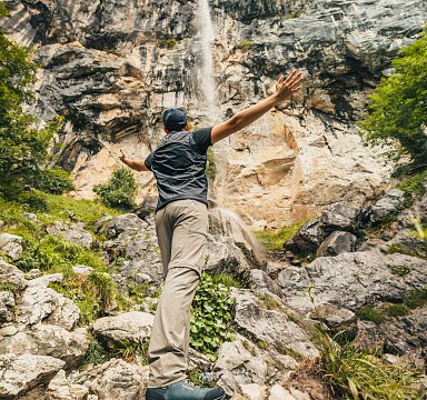 Ein Mensch steht mit ausgestreckten Armen vor einem Wasserfall, umgeben von Felsen und dichter Vegetation, und blickt nach oben zur Quelle.