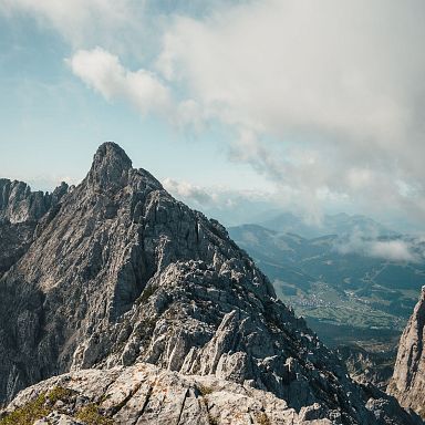 Majestätisches Bergmassiv mit spitzen Gipfeln unter leicht bewölktem Himmel, im Hintergrund sanfte Täler und ein weitläufiger Blick auf die Alpenlandschaft.