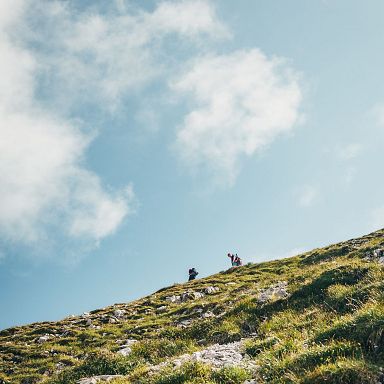 Wanderer erklimmen einen grasbewachsenen Hügel unter blauem Himmel mit Wolken, symbolisierend Freiheit und Abenteuer in der Natur.