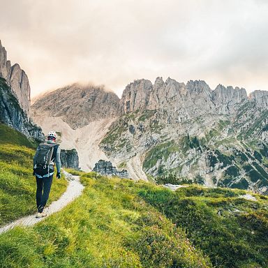 Ein Wanderer mit Rucksack geht auf einem schmalen Pfad durch eine alpine Landschaft mit grünen Wiesen im Vordergrund und steilen Felsformationen im Hintergrund.