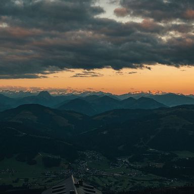 Blick auf eine Berglandschaft bei Sonnenuntergang, mit schneebedeckten Gipfeln am Horizont und einem dramatischen Himmel voller dunkler Wolken.