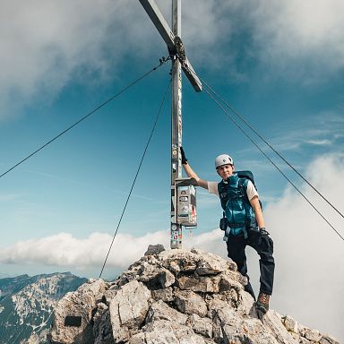 Ein Bergsteiger steht neben einem Gipfelkreuz auf einem felsigen Gipfel, umgeben von einer weiten Berglandschaft und ein paar Wolken am Himmel.