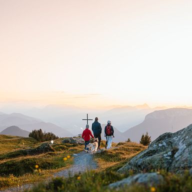 Three people hike a mountain path at sunset in the Wilder Kaiser, surrounded by rugged terrain and overlooking valleys. A cross stands on the trail ahead.