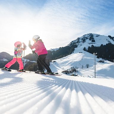 Ein Kind und ein Erwachsener in bunter Skikleidung fahren bei Sonnenschein auf einer schneebedeckten Piste in den Bergen.