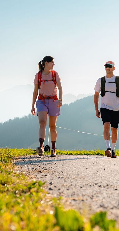 Zwei Personen wandern auf einem sonnigen Pfad mit Alpenlandschaft im Hintergrund und tragen sportliche Kleidung. Der Himmel ist klar und die Aussicht ist beeindruckend.