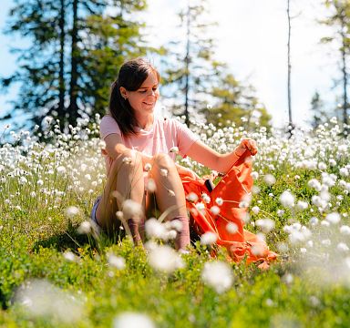 Frau sitzt lächelnd auf einer Blumenwiese, umgeben von weißen Blüten, im Sonnenlicht. Sie hält ein orangefarbenes Tuch in der Hand. Im Hintergrund sind Bäume zu sehen.