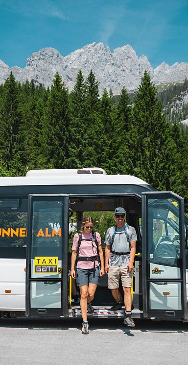 Hiking bus with people stepping out in front of the majestic Wilder Kaiser mountains, surrounded by lush greenery and rugged peaks under a clear blue sky.