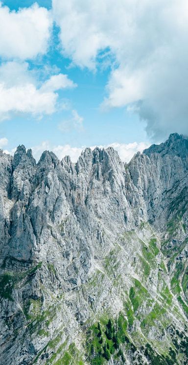 Steile, zerklüftete Berggipfel des Wilden Kaisers unter blauem Himmel mit Wolken. Die Felsen sind teils bewachsen, teils kahl und strahlen Erhabenheit aus.