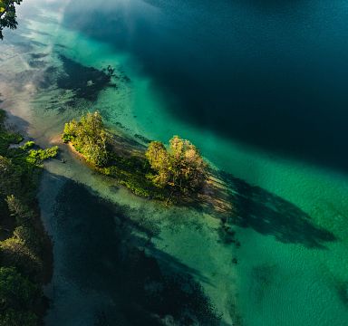 Luftaufnahme eines klaren Sees mit grün-blauem Wasser, bewaldetem Ufer und kleinen Inseln. Sonnenlicht wirft Schatten auf die Wasseroberfläche.