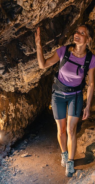 Eine Frau in sportlicher Kleidung wandert durch einen felsigen Höhlengang. Sie berührt die Decke und lächelt, umgeben von warm beleuchtetem Gestein.