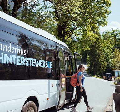 A woman with a backpack boards a hiking bus to Hintersteiner See in the Wilder Kaiser region. The bus is parked on a road surrounded by green trees and a blue sky.