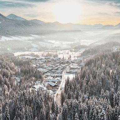 A serene winter scene at Wilder Kaiser with snow-covered trees and mountains framing a peaceful village under a warm sunrise.