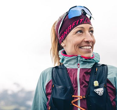 Person in Outdoor-Kleidung und Stirnband, lächelnd, mit einem nebligen Bergpanorama im Hintergrund. Die Stimmung ist sportlich und abenteuerlich.