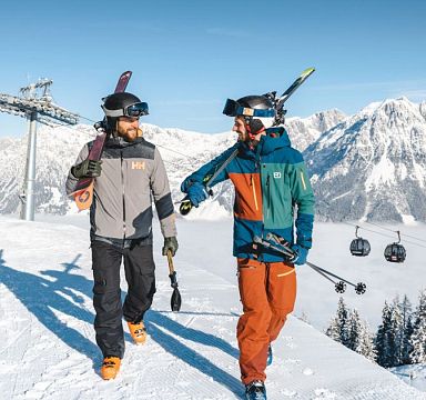 Zwei Herren tragen Ski über der Schulter im Hintergrund Bergbahn und Gipfel