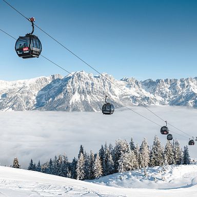 Snowy mountains of Wilder Kaiser with cable cars and cloud-covered valley, trees dusted in snow under a clear blue sky, offering a picturesque winter view.