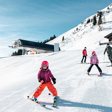 Familie fährt bei strahlend blauem Himmel auf frisch präparierten Pisten Ski. Ein modernes Liftgebäude und eine rustikale Holzhütte sind im Hintergrund zu sehen.