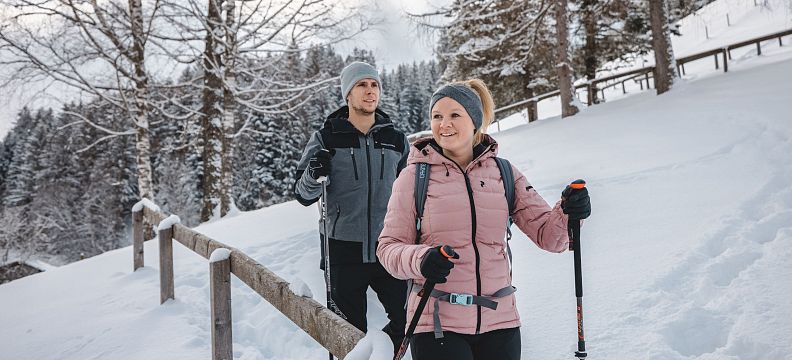 Two hikers in winter clothing walk along a snowy path in Wilder Kaiser, with trees and snow-covered ground, surrounded by a peaceful mountain landscape.