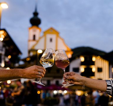 Two people clinking wine glasses in front of lit buildings in an alpine setting, evening atmosphere in the Wilder Kaiser region, symbolizing celebration.