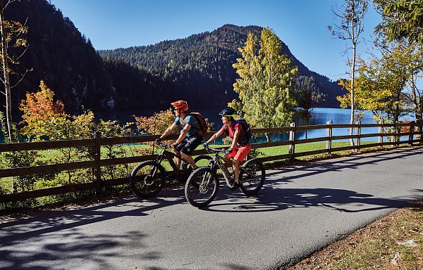 Two cyclists ride on a paved path beside a tranquil lake, with autumn trees and mountains in the background, creating a picturesque scene.
