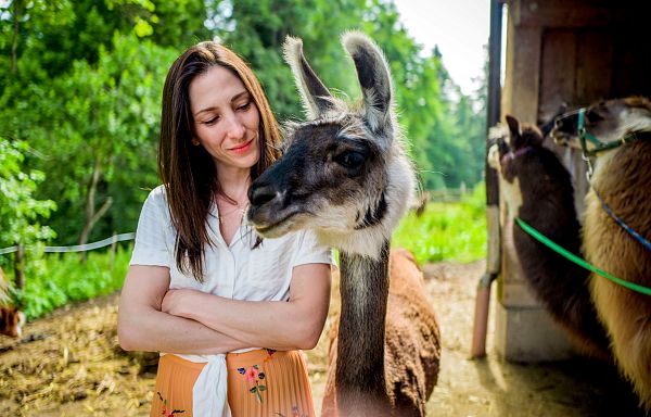 Frau steht lächelnd mit einem Lama in einer ländlichen, grünen Umgebung. Im Hintergrund sind weitere Lamas zu sehen.