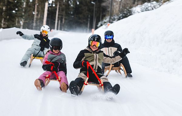 A group of people sledding down a snow-covered hillside, surrounded by trees, in the Wilder Kaiser region of Tirol. They are dressed in winter gear.