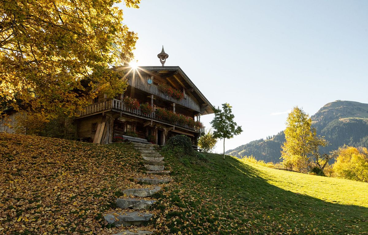 Almhütte im Herbst, umgeben von buntem Laub und Bergen im Hintergrund, unter strahlendem Sonnenschein und klarem Himmel.