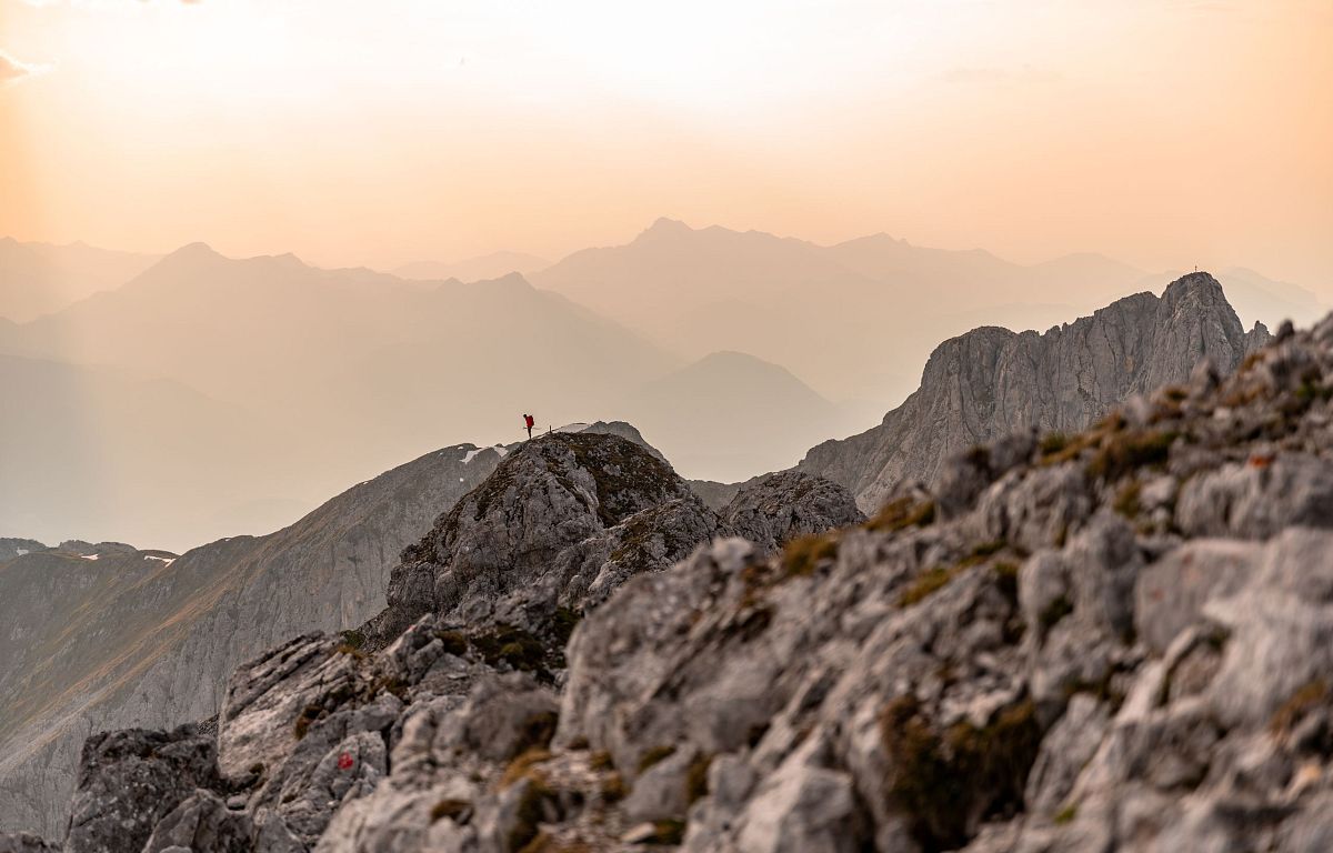 Ein einsamer Wanderer steht auf einem felsigen Gipfel und blickt in die Ferne. Der Himmel ist in warmen Orange- und Gelbtönen der Abenddämmerung gefärbt.