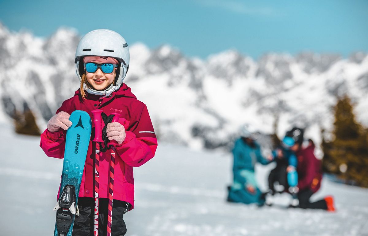 A child in red ski gear smiles on the slopes of Wilder Kaiser, with snowy mountains and other skiers in the background.