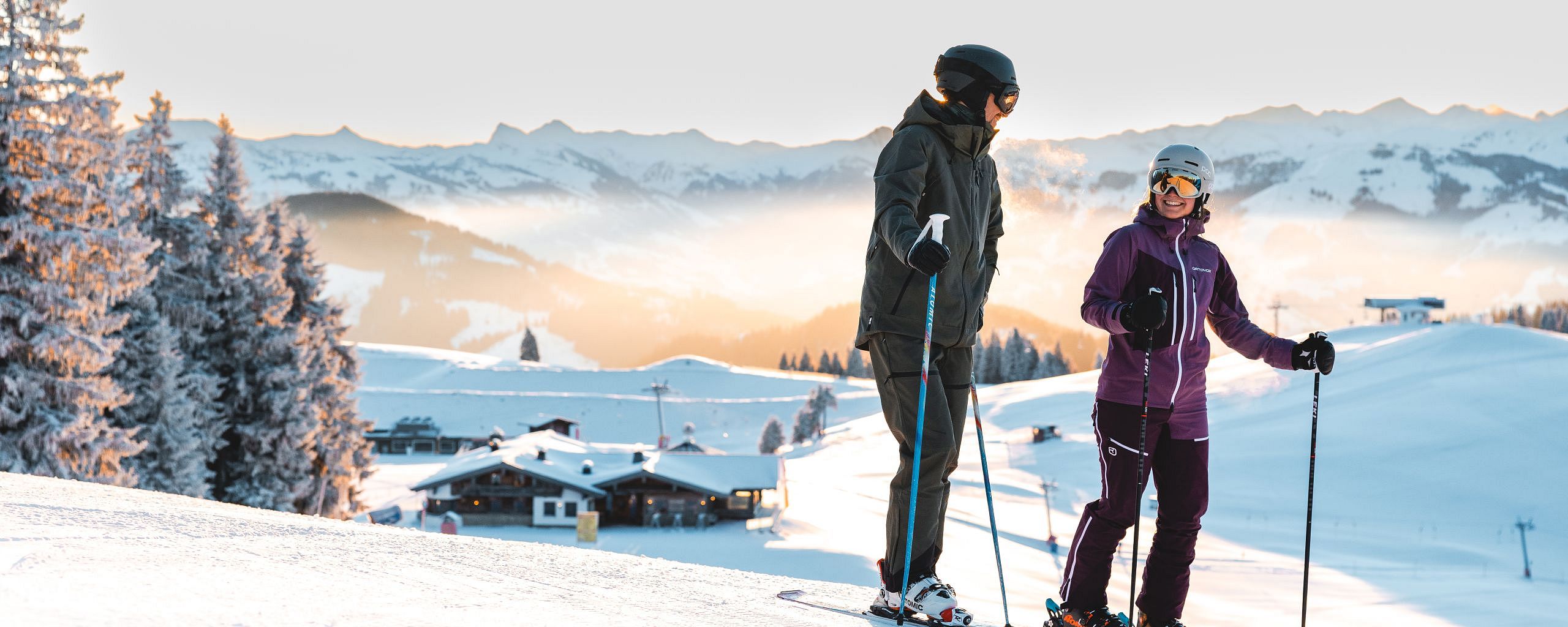 Two skiers pause on a snowy slope with majestic mountains and a sunlit sky in the background at Wilder Kaiser, surrounded by snow-covered trees and a cozy cabin.