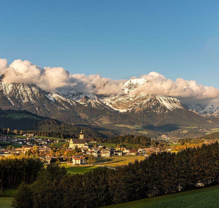 Landschaft im Herbst mit einem Dorf im Tal, umgeben von schneebedeckten Bergen unter bewölktem Himmel. Die Bäume im Vordergrund sind teils grün, teils gelb.