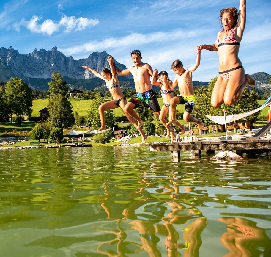 Vier Personen springen von einem Steg in einen See, im Hintergrund beeindruckende Bergkulisse des Wilden Kaisers, grüner Wald und blauer Himmel.