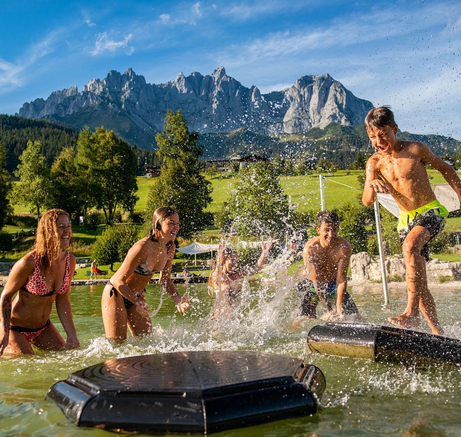 Kinder spielen fröhlich im Wasser an einem sonnigen Tag, im Hintergrund erhebt sich eindrucksvoll das Bergmassiv des Wilden Kaisers in Tirol.