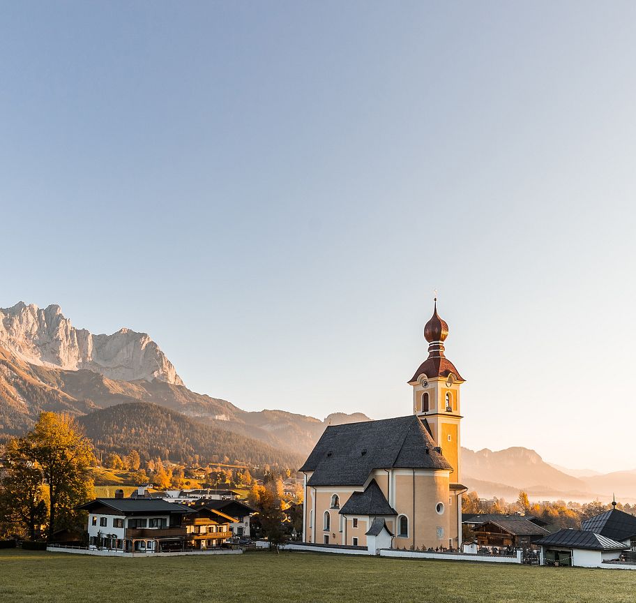 Eine malerische Kirche mit roter Kuppel liegt vor dem beeindruckenden Bergmassiv des Wilden Kaisers, umgeben von herbstlichen Bäumen und klarem Himmel.