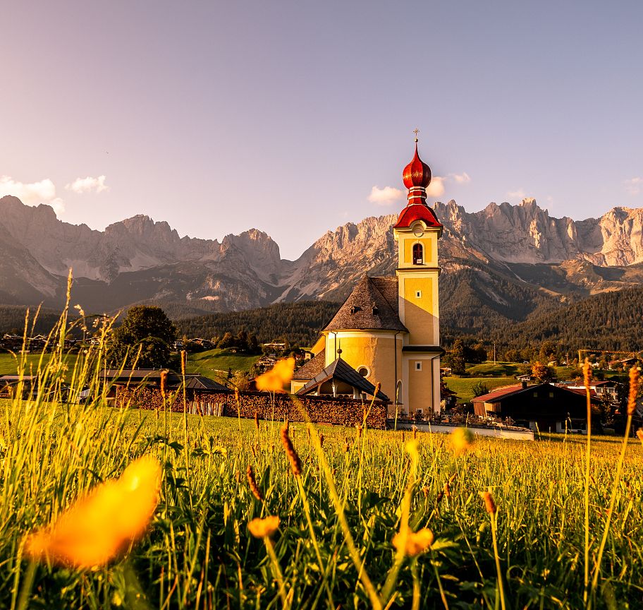 A small church with a red dome in a green meadow, surrounded by mountains of the Wilder Kaiser under a clear, sunny sky.