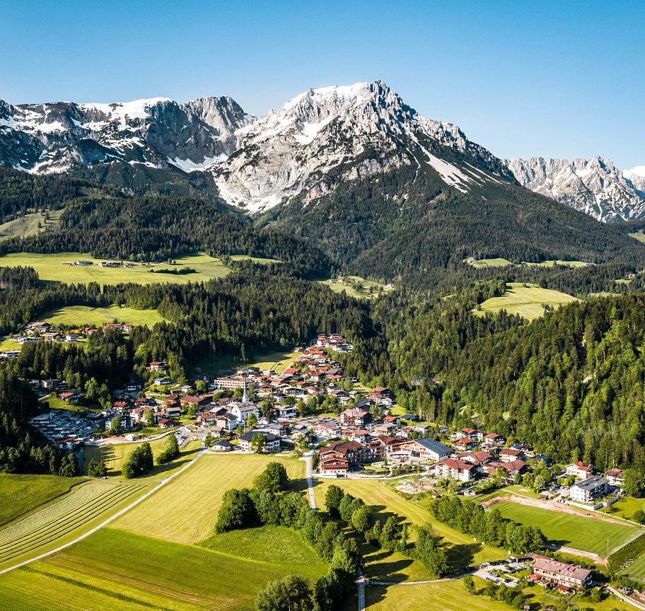 Luftaufnahme eines malerischen Alpendorfs umgeben von grünen Wiesen und dichten Wäldern, mit einem imposanten Bergmassiv im Hintergrund unter klarem, blauem Himmel.