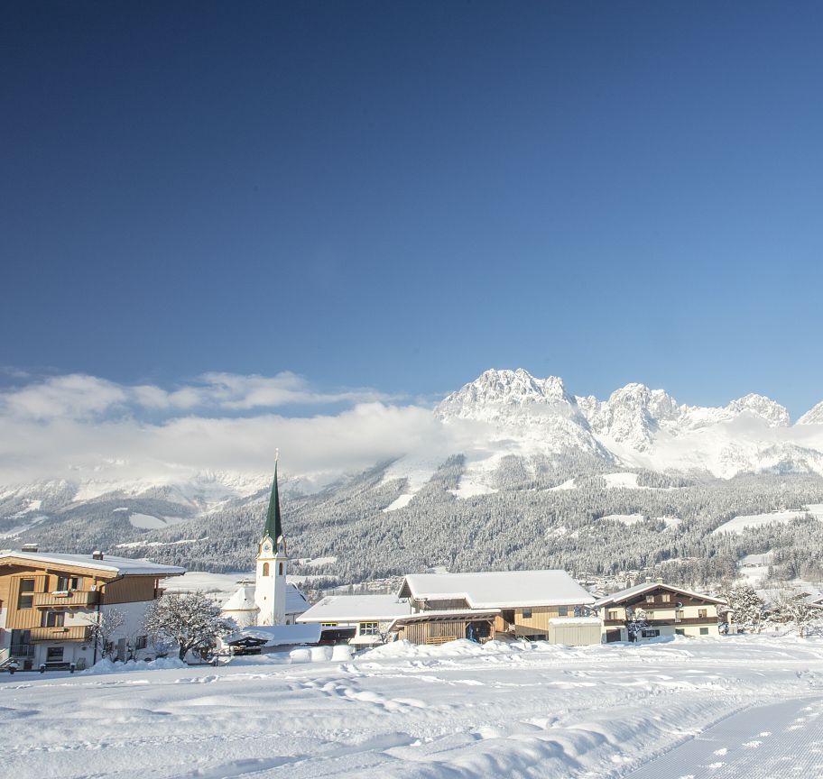 Winterliches Panorama in den Alpen mit schneebedeckten Bergen, einem Dorf mit Kirche und blauen Himmel. Perfekte Szene für Winterurlaub und Skifahren.