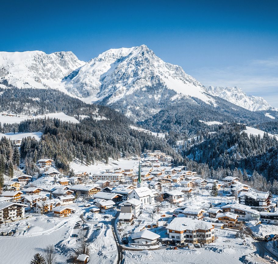 Snow-covered village with the majestic Wilder Kaiser mountains in the background, under a clear blue sky, capturing the essence of a peaceful winter day.