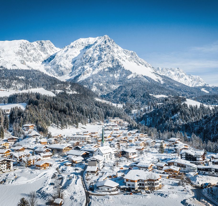 Snow-covered village with the majestic Wilder Kaiser mountains in the background, under a clear blue sky, capturing the essence of a peaceful winter day.