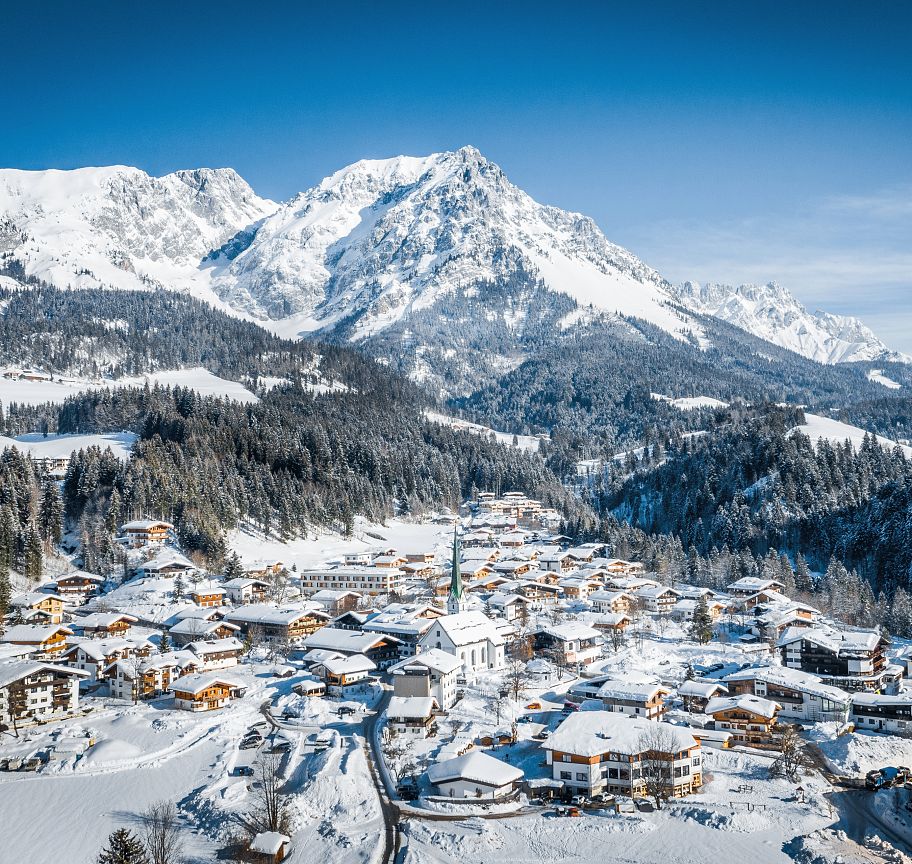 Snow-covered village with the majestic Wilder Kaiser mountains in the background, under a clear blue sky, capturing the essence of a peaceful winter day.