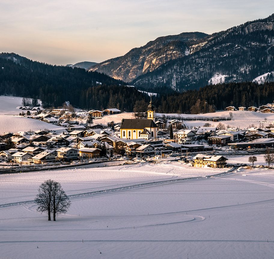 A snow-covered village nestled in the Wilder Kaiser region, with mountains in the background and warm sunlight illuminating the landscape.