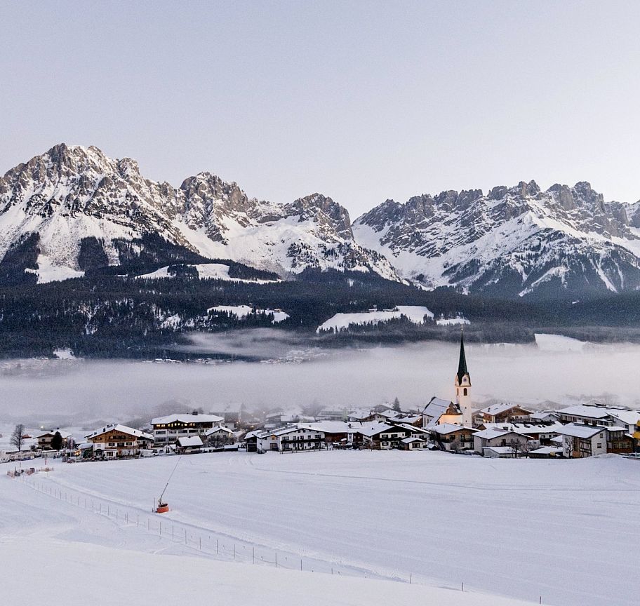 Snow-covered Wilder Kaiser mountains with a village below, partially covered in morning mist.