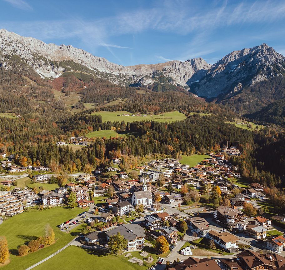 Luftaufnahme eines Dorfes in den Alpen mit traditionellen Häusern, umgeben von grünen Wiesen und Wäldern. Im Hintergrund erhebt sich ein imposantes Bergmassiv.