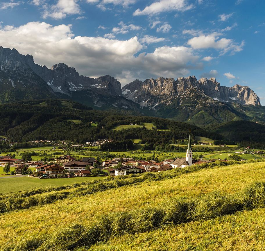 Panorama der Region Wilder Kaiser mit majestätischem Bergmassiv im Hintergrund, grünen Wiesen im Vordergrund und einem Dorf mit einer markanten Kirche.