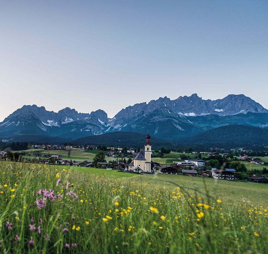 Ein blühendes Wiesenfeld mit bunten Blumen im Vordergrund, dahinter ein malerisches Dorf mit einer Kirche, vor den majestätischen Bergen des Wilden Kaisers.