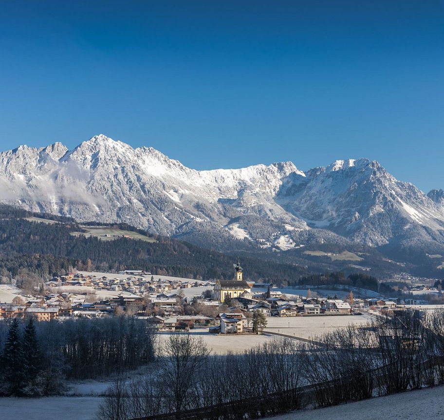 Panorama des Wilder Kaiser Gebirges im Winter. Verschneite Berggipfel erheben sich über ein malerisches Tal mit einem kleinen Dorf und unberührter Natur.