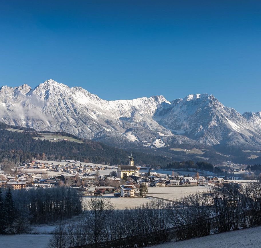 Snow-covered peaks of the Wilder Kaiser under a blue sky, with a small village in the valley. The landscape is bright and serene, capturing winter's essence.